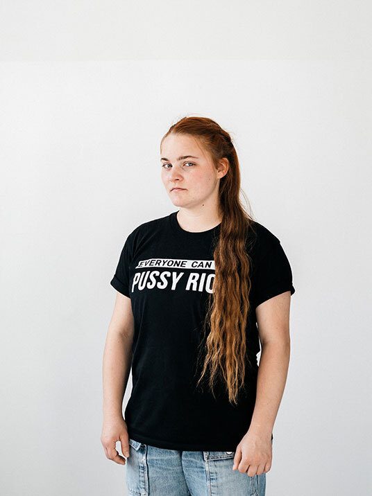 Eine junge Frau mit langen, roten Haaren steht vor einem weißen Hintergrund. Sie trägt ein schwarzes T-Shirt mit dem Aufdruck "EVERYONE CAN PUSSY RIOT" und Jeans. Ihr Blick ist direkt und wirkt entschlossen.