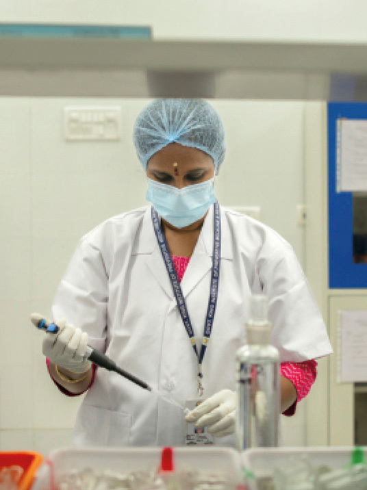 Eine Frau in einem Labor arbeitet konzentriert mit einer Pipette an einer Probe. Sie trägt einen Haarschopfschutz, eine Mundmaske und einen weißen Laborkittel. Sie wirkt professionell und engagiert.
