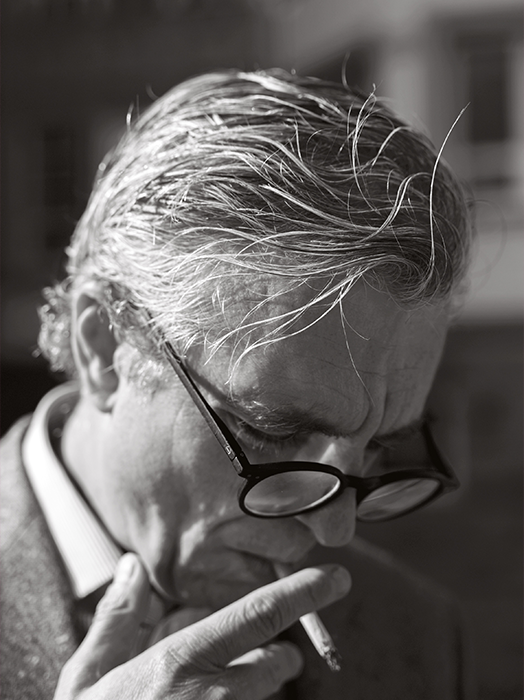 Das Bild ist eine Schwarz-Weiß-Fotografie eines Mannes mit grauem Haar, der eine Brille trägt und eine Zigarette raucht. Der Mann ist in der unteren Hälfte des Bildes positioniert, wobei sein Kopf leicht nach links geneigt ist. Der Hintergrund ist unscharf und lenkt die Aufmerksamkeit auf den Mann und seine Zigarette. Das Bild enthält keinen erkennbaren Text.
